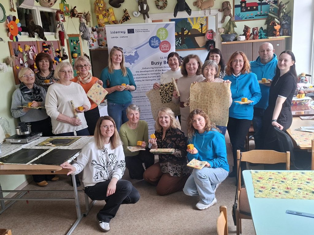 Group of diverse participants engaged in a crafting workshop, showcasing handmade products, with a focus on community art and creativity. The setting features colorful decorations and a banner promoting the "Busy Bee" project, highlighting collaboration between Latvia and Lithuania.
