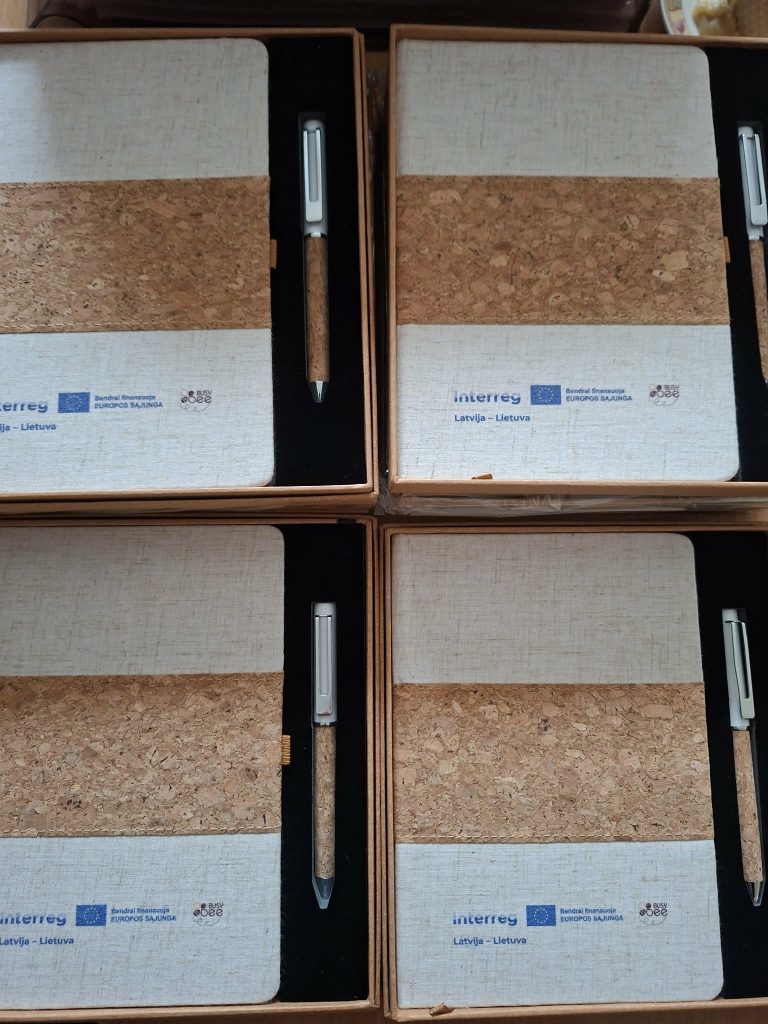 Eco-friendly notebooks with cork accents and matching pens, featuring branding for the Interreg project between Latvia and Lithuania, arranged neatly in boxes.