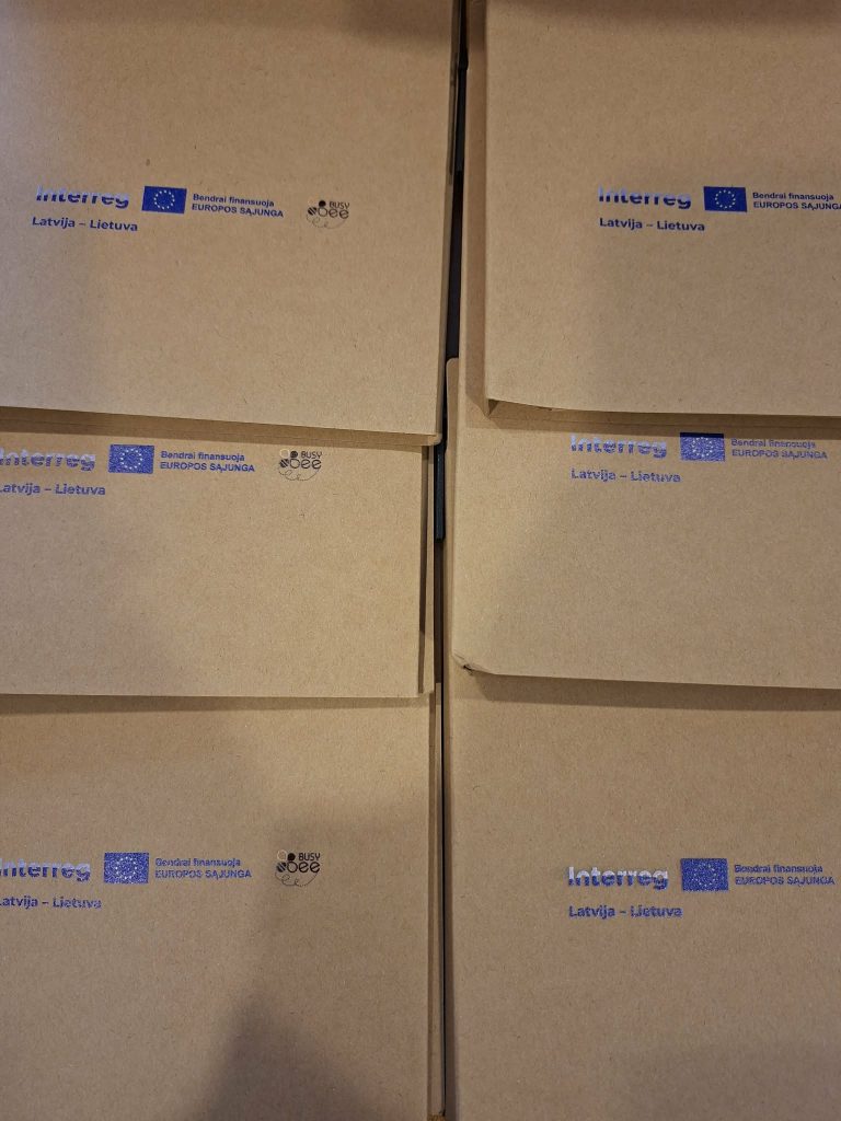 Brown envelopes featuring the Interreg logo and text "Latvija - Lietuva," representing the EU-funded cooperation program between Latvia and Lithuania.