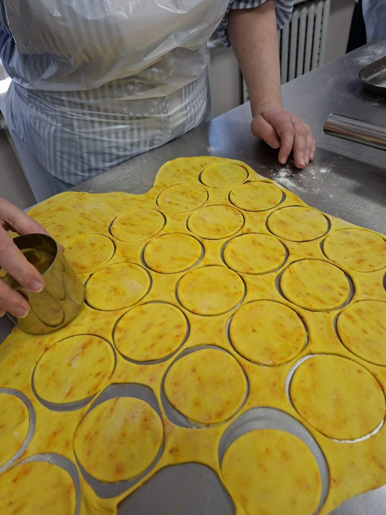 Person cutting circular shapes from yellow dough on a stainless steel countertop, surrounded by flour, preparing for baking or cooking.