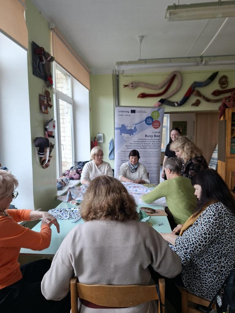 Group of women participating in a crafting workshop, focused on textile art, in a bright room with a banner for the Busy Bee project. Various colorful fabrics are spread out on the table, promoting creativity and collaboration among participants.