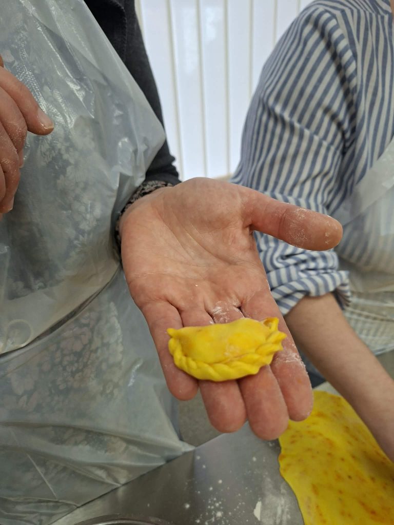Person holding a freshly made yellow ravioli in a kitchen setting, with flour on the table and a second person in the background wearing an apron.