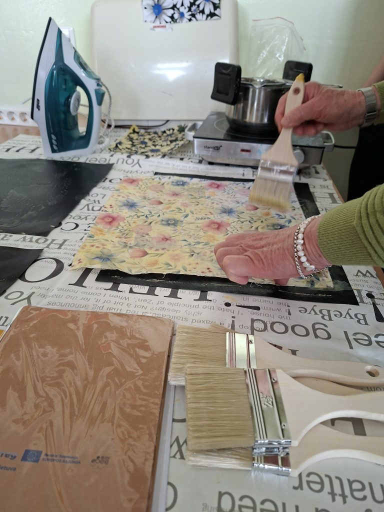 An elderly person applying decoupage technique with a brush on floral-patterned paper, set on a craft table with paintbrushes, an iron, and a stovetop in the background.
