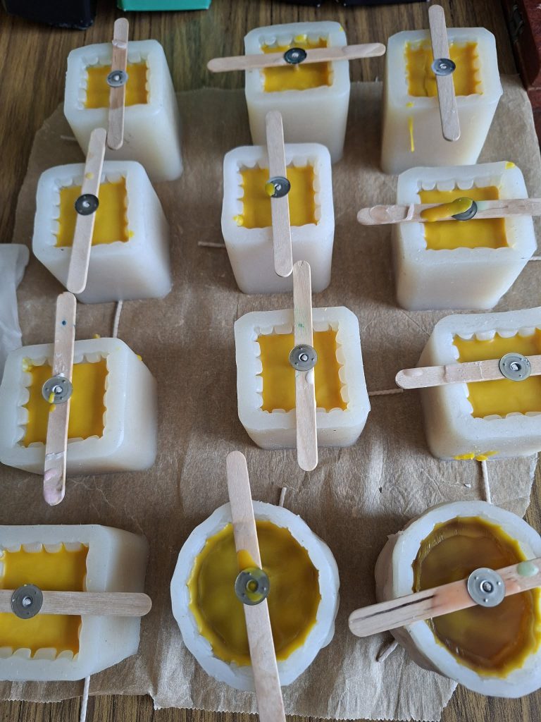 Handmade candles in silicone molds with wooden wicks, featuring a bright yellow wax filling. The molds are placed on a brown paper surface, showcasing various shapes and sizes of the candles.
