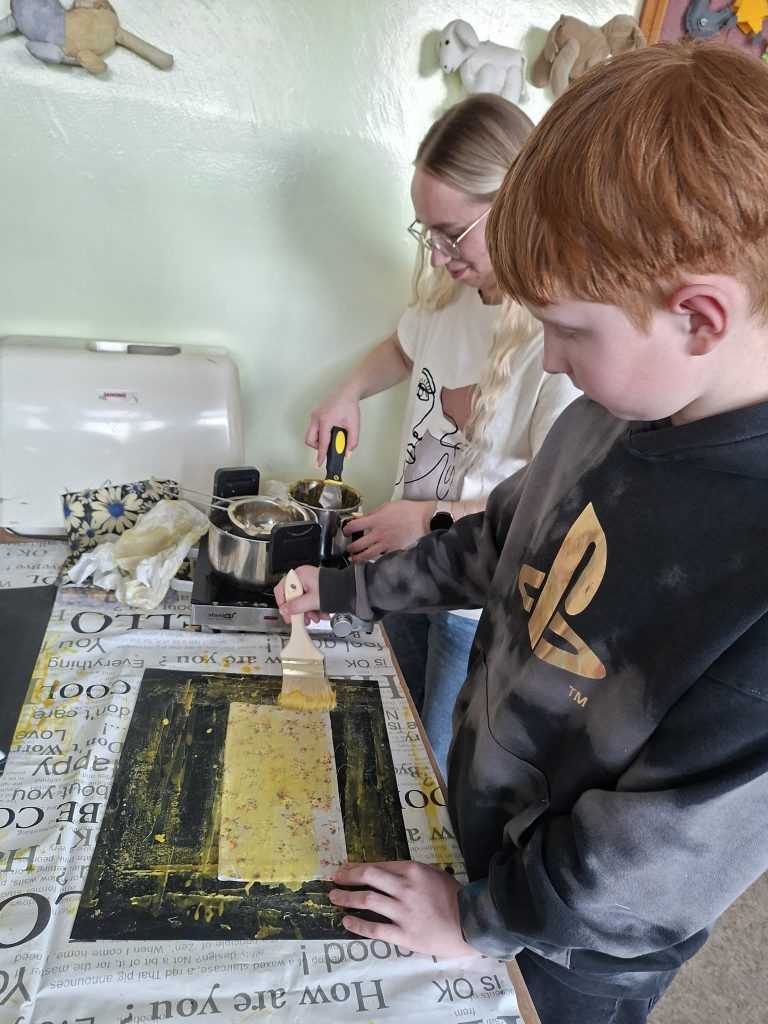 A child and an adult engaged in a creative painting activity, with the child using a brush to apply yellow paint to a canvas on a table covered with a patterned tablecloth. The adult is mixing materials in a bowl, illustrating a hands-on art project.