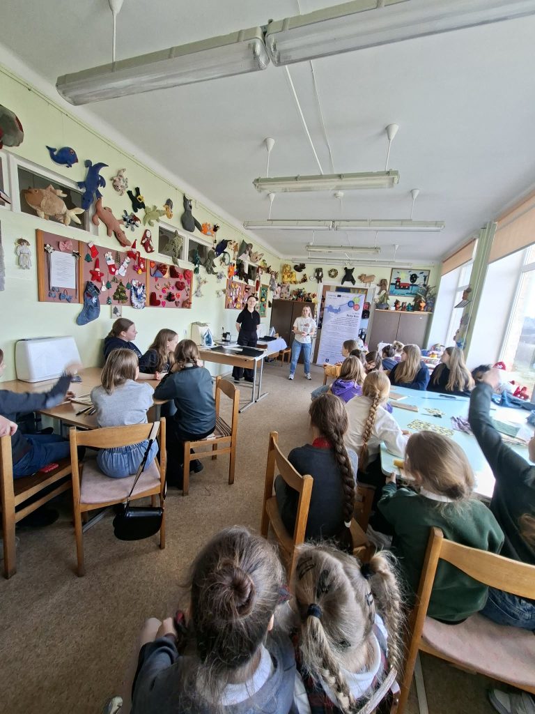 Classroom filled with children engaged in an art activity, with colorful artwork displayed on the walls, and instructors presenting at the front.