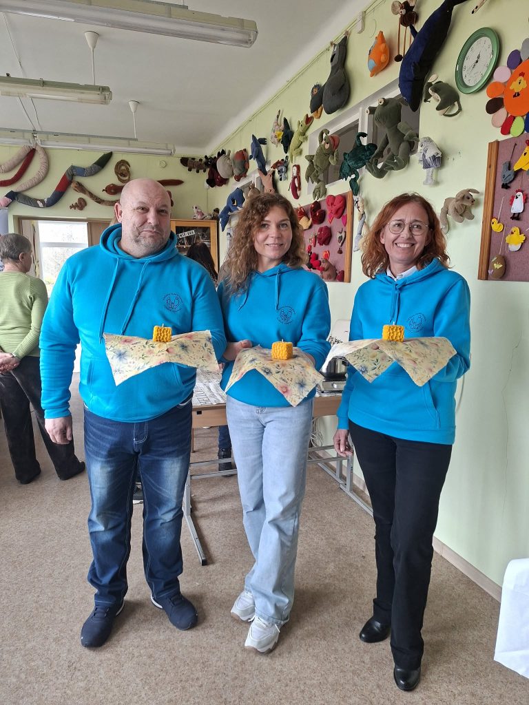 Three individuals wearing matching blue hoodies stand together indoors, each holding a decorated fabric wrap with a yellow item on top. The backdrop features colorful crafts displayed on the walls, indicating a creative environment.