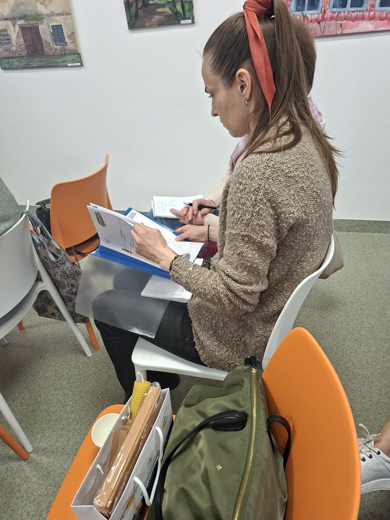 Woman sitting in a chair, reviewing documents and taking notes during a meeting or workshop. She has long hair tied with a ribbon, wearing a beige sweater, and is surrounded by colorful chairs and artwork on the walls. A green bag and a box of supplies are placed beside her.