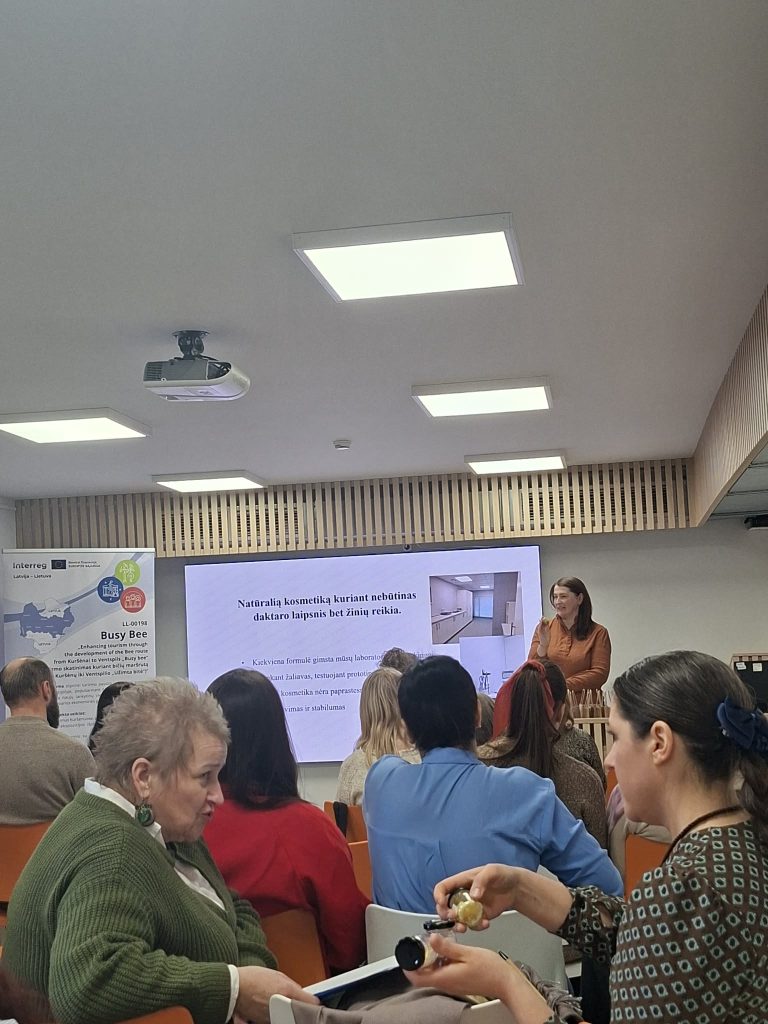 Audience at a seminar on natural cosmetics, featuring a speaker presenting information on cosmetic formulation. The presentation includes visuals related to laboratory practices. Attendees are engaged, with some taking notes and looking at materials.