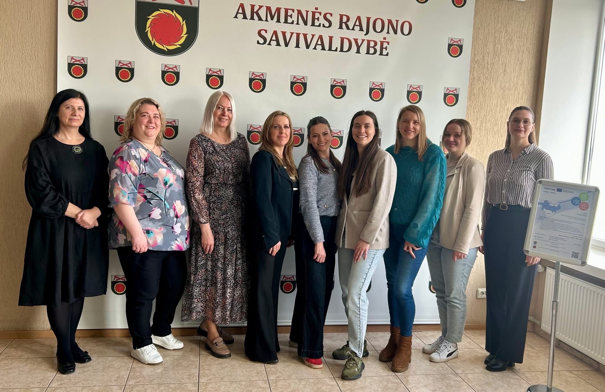 Group of professional women posing in front of the Akmenės Rajono Savivaldybė sign, showcasing teamwork and community engagement in a formal setting.