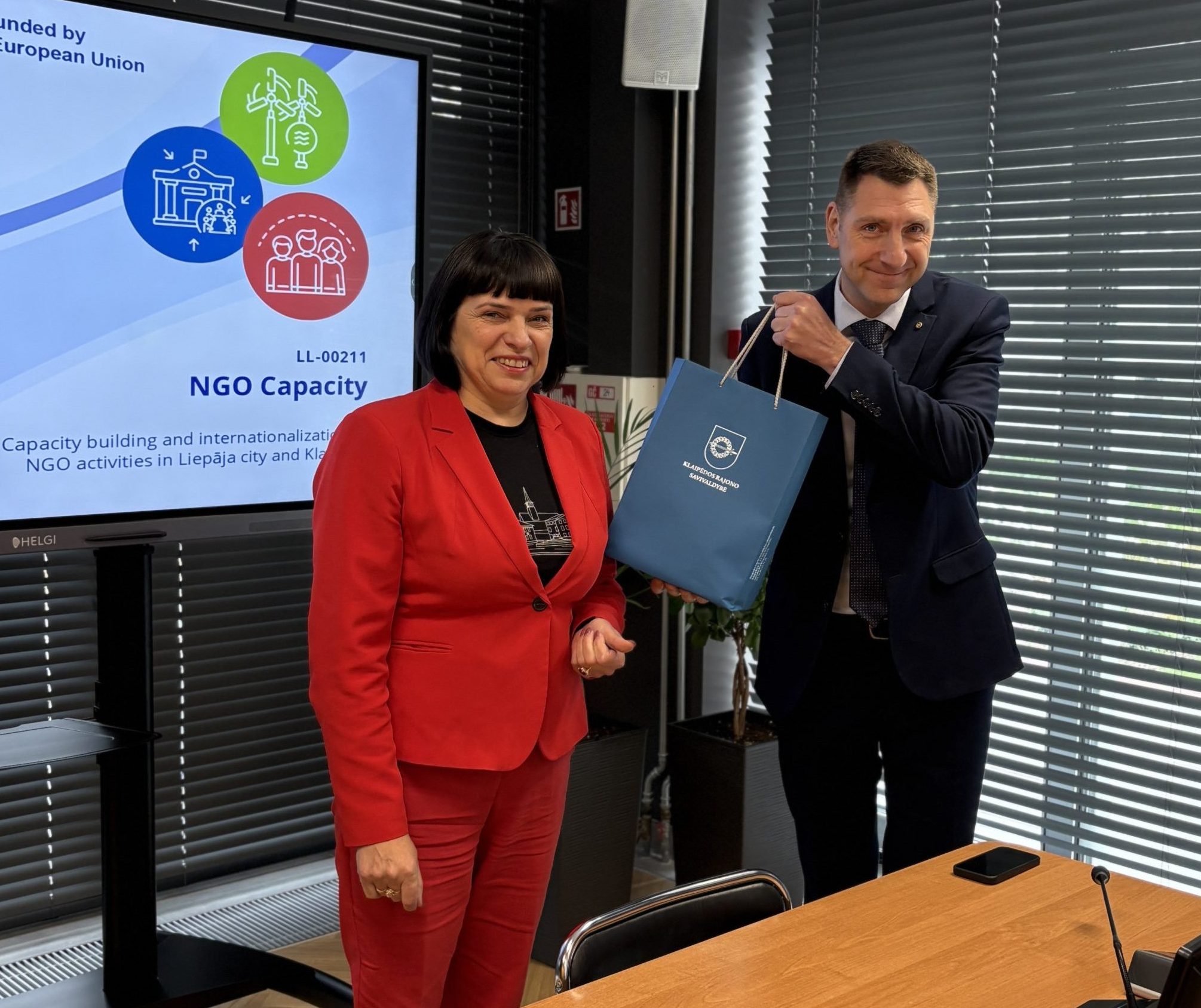 Two professionals engage in a meeting, with one presenting a blue bag while the other smiles. In the background, a screen displays information about an NGO capacity-building project co-funded by the European Union, focusing on enhancing NGO activities in Liepāja city and Kuldīga. The setting features a modern conference room with wooden tables and large windows.