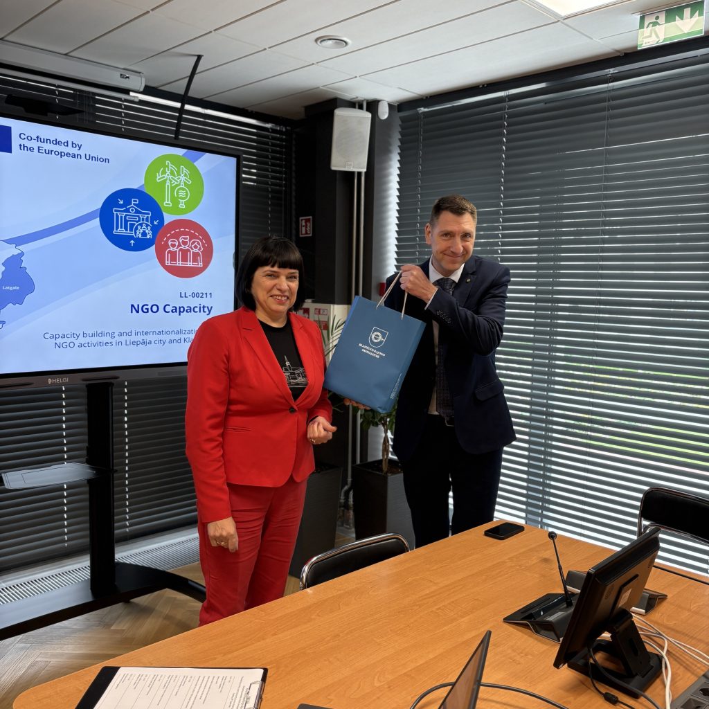 Two professionals engage in a discussion at a conference room, with one presenting a blue gift bag and the other wearing a red suit. A screen in the background displays information about an NGO capacity building project funded by the European Union, highlighting activities in Liepāja city. The setting includes a wooden table and modern office decor, emphasizing collaboration in NGO initiatives.