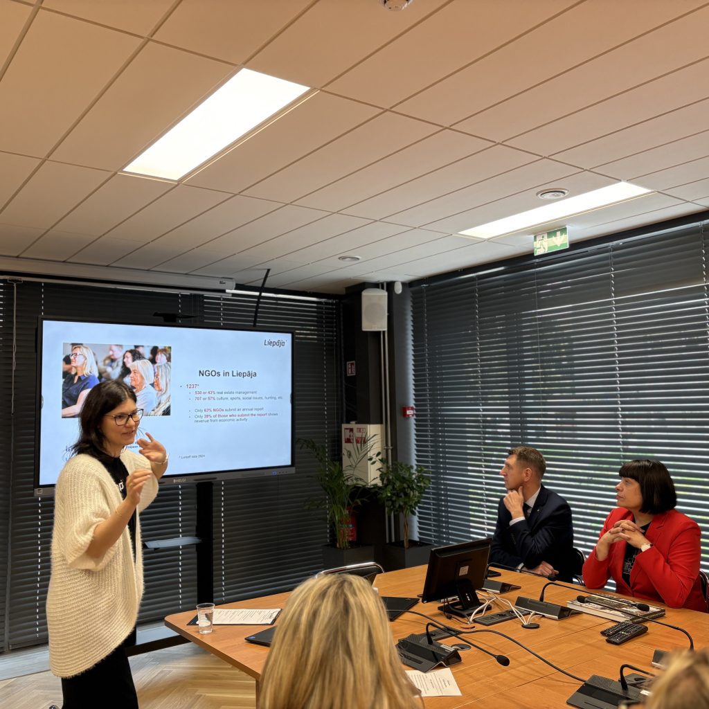 A woman presents information on NGOs in Liepāja during a meeting, with colleagues attentively listening. A large screen displays key statistics and insights about local NGOs, highlighting community engagement and social management initiatives.