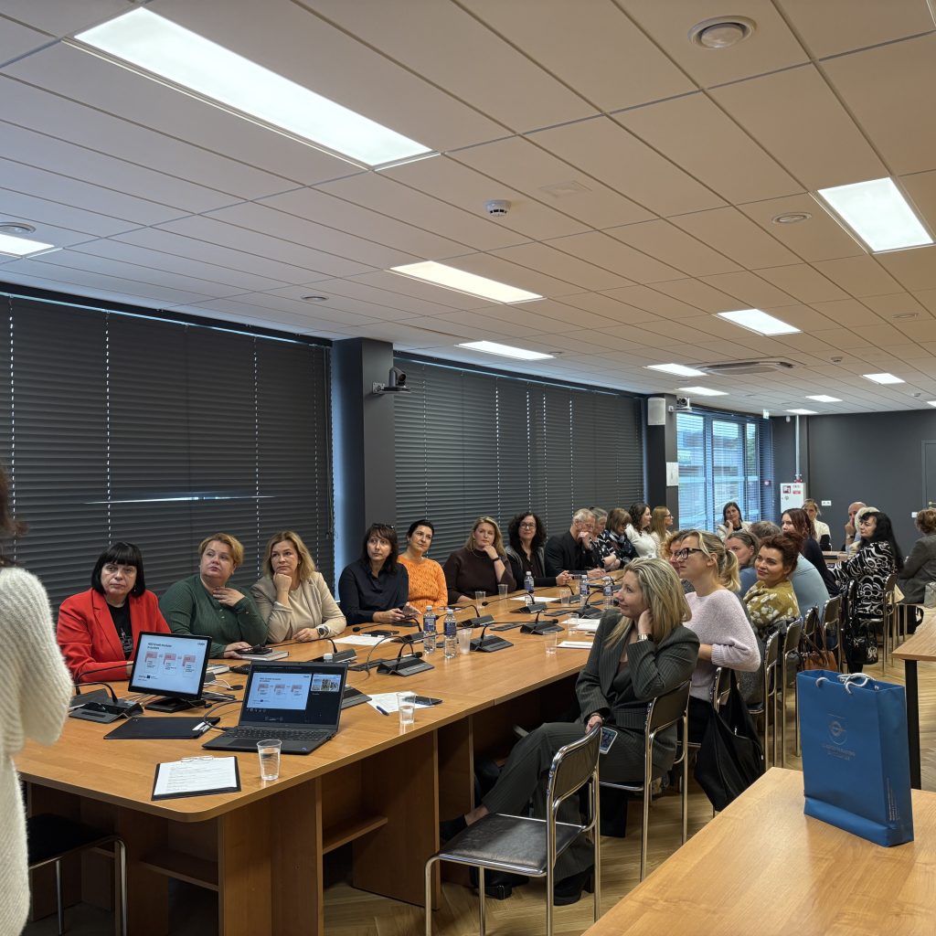 A group of women attending a professional seminar at a conference table, engaged in discussion. Laptops and notepads are visible, with a presenter speaking at the front of the room. The setting features modern decor and large windows with blinds.