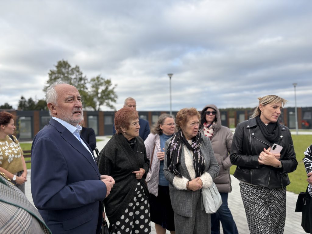 Group of diverse adults attentively listening to a speaker outdoors, with a cloudy sky in the background. The scene captures a moment of engagement during a community event or gathering, showcasing individuals in various attire, including formal and casual clothing.