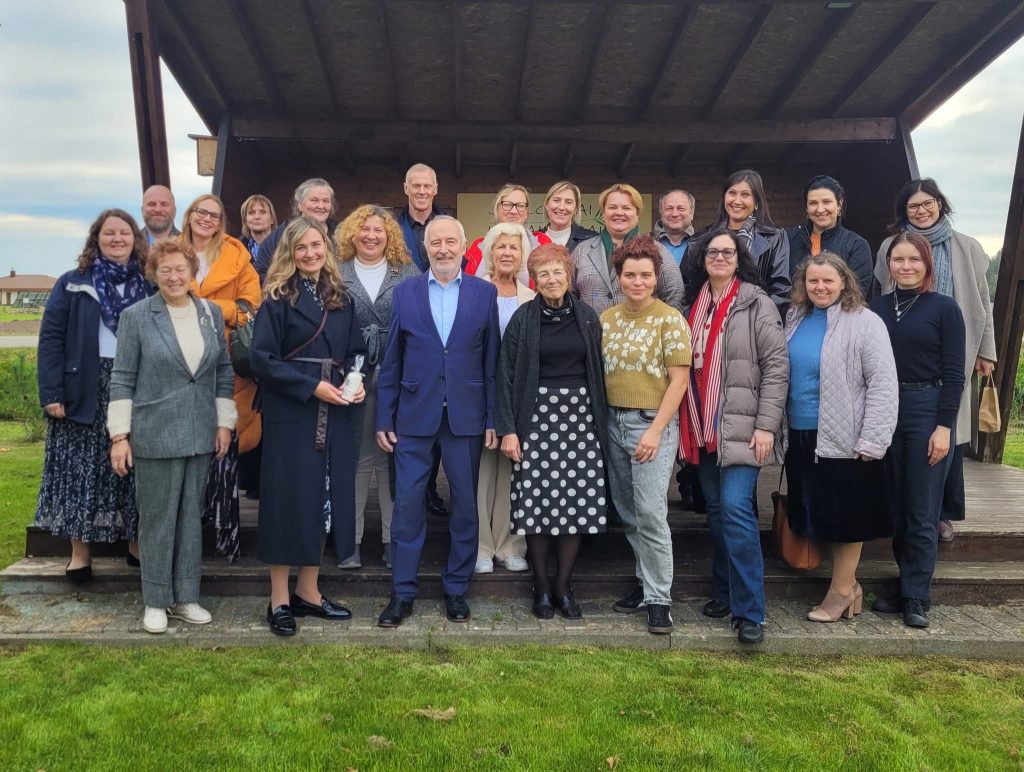 Group photo of diverse professionals gathered outdoors in front of a wooden structure, showcasing a mix of formal and casual attire, smiling and engaged in a community event.
