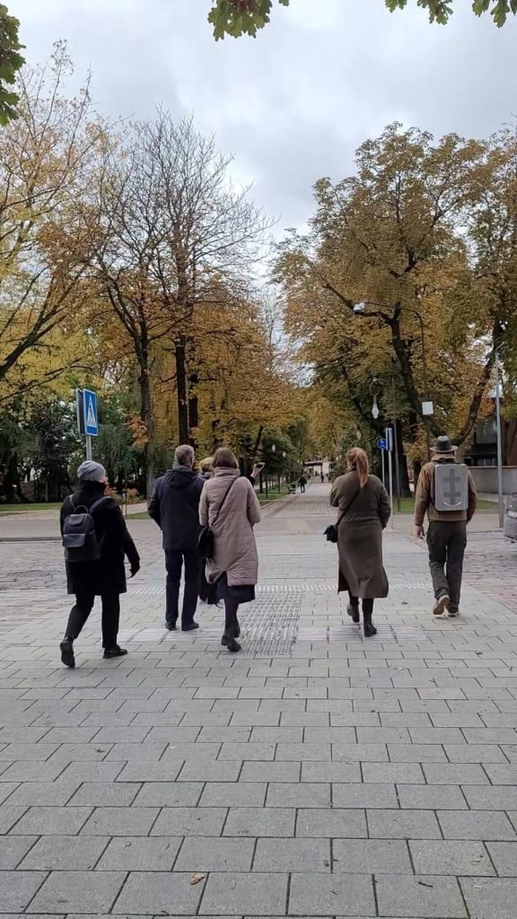 Group of five people walking along a tree-lined street in autumn, showcasing colorful foliage and a cloudy sky. The scene captures a peaceful urban environment, ideal for leisurely strolls.