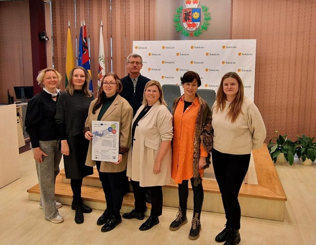 Group of professionals standing together in a municipal office in Šiauliai, Lithuania, holding a document. The setting features the city’s flags and a logo backdrop, highlighting collaboration and community engagement.