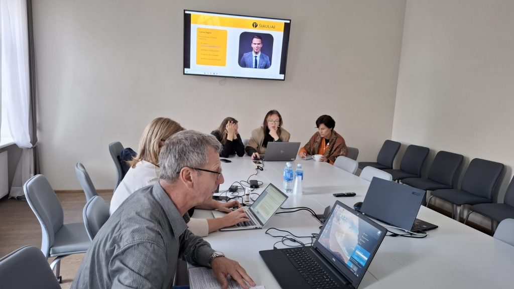 Team meeting in a modern conference room with laptops and a presentation screen displaying a speaker. Participants, including four women and one man, are engaged in discussion and collaboration. Water bottles and notepads are visible on the table, emphasizing a professional environment.