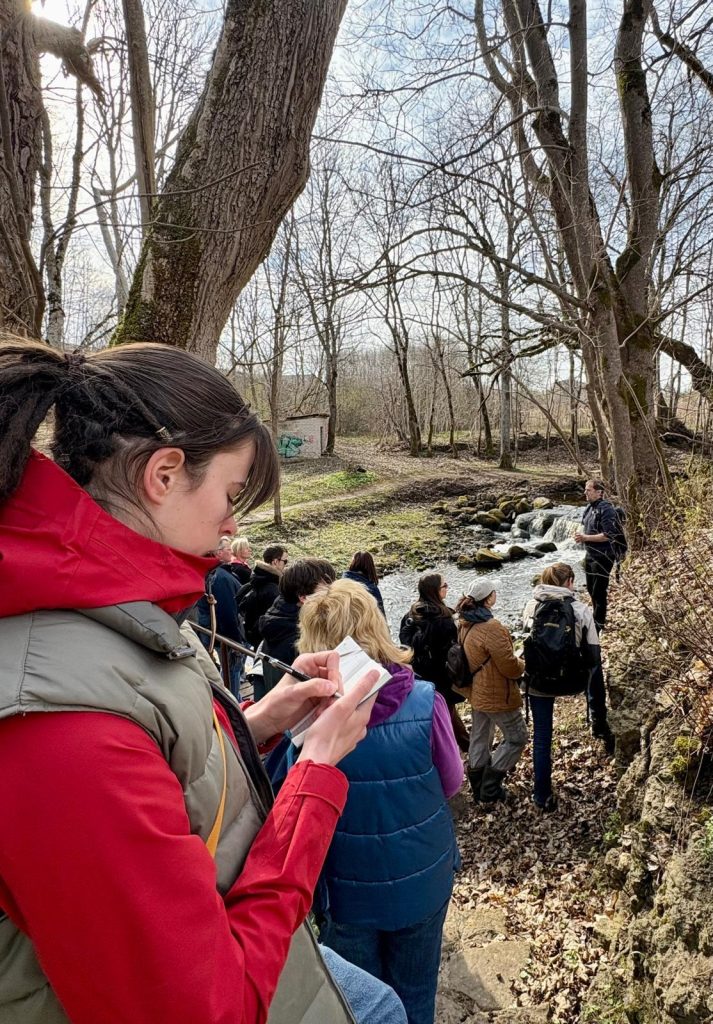 A group of people gathered near a stream in a forested area, with one individual taking notes. The scene captures a nature-focused educational activity, showcasing trees in early spring and participants engaged in learning.