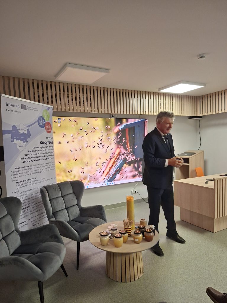Speaker presenting at a conference on beekeeping with a display of honey jars and a background image of bees. The setting features modern furniture and promotional materials related to the Busy Bees project, highlighting the importance of bee conservation and tourism development in Latvia and Lithuania. Speaker presenting at a conference on beekeeping with a display of honey jars and a background image of bees. The setting features modern furniture and promotional materials related to the Busy Bees project, highlighting the importance of bee conservation and tourism development in Latvia and Lithuania.