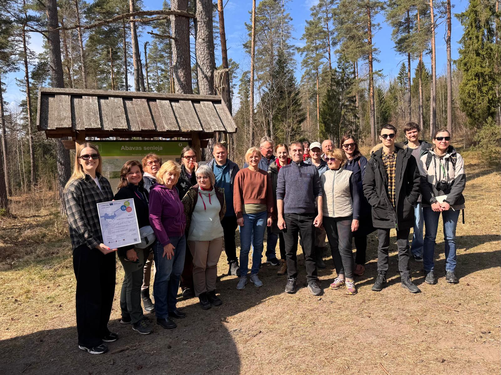 Group of individuals gathered at Abavas senleja in a forest setting, celebrating an event. The scene features a mix of people holding a certificate, surrounded by tall trees and a clear blue sky, emphasizing outdoor community engagement and nature appreciation.