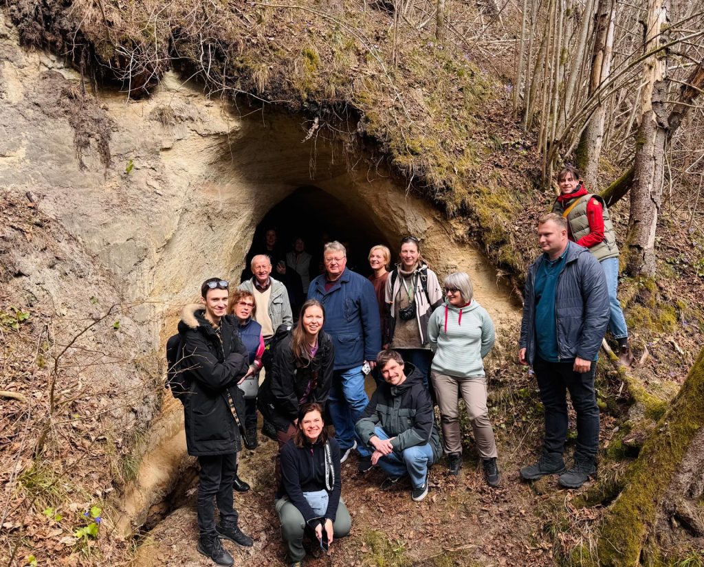 Group of hikers gathered at the entrance of a cave in a forested area, surrounded by trees and natural foliage, showcasing a springtime outing and adventure in nature.