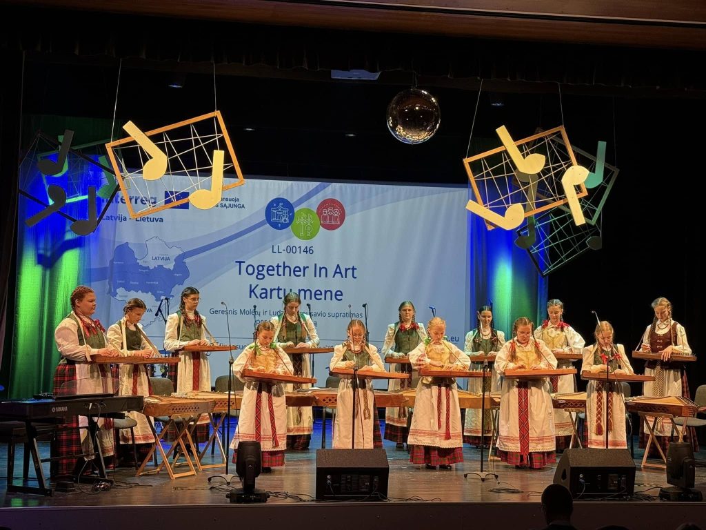 A group of performers in traditional Latvian attire playing musical instruments on stage at the "Together In Art" event, with a backdrop featuring the event's logo and decorative musical notes.