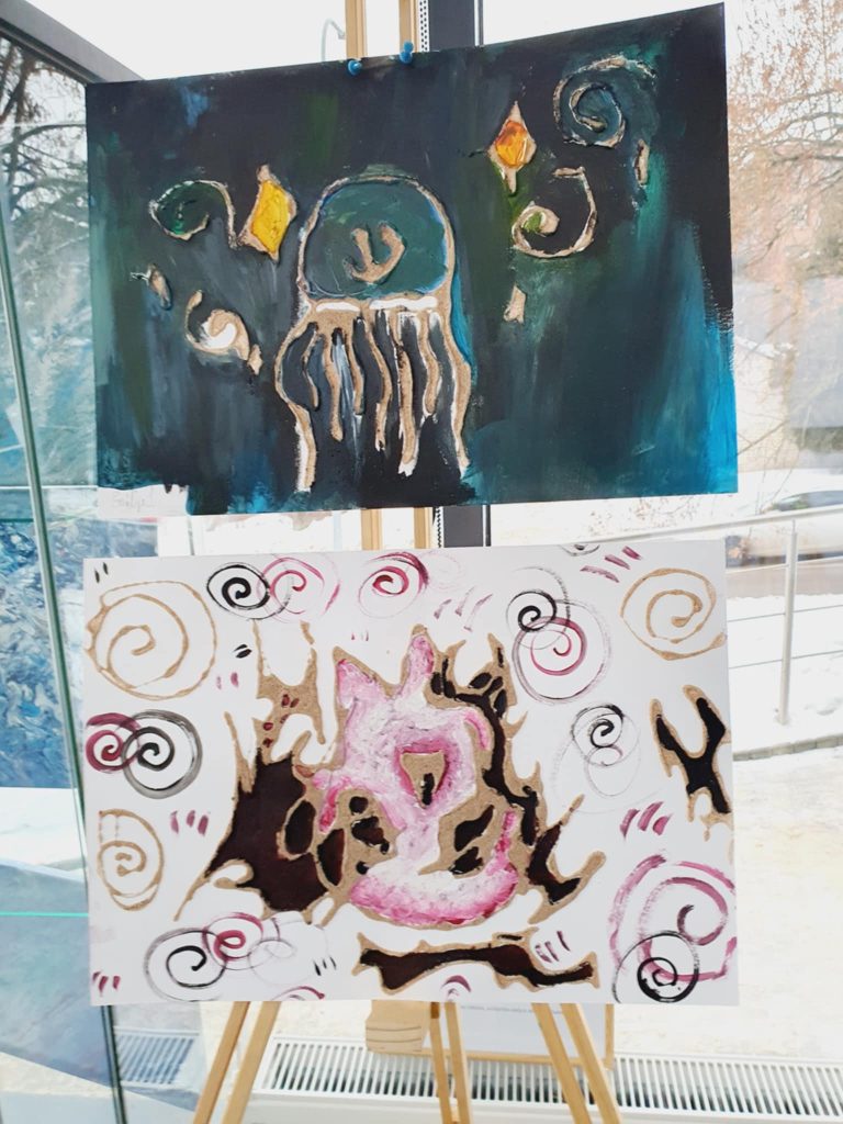 Two colorful abstract paintings displayed on an easel. The top artwork features a jellyfish with swirling patterns and vibrant colors against a dark background. The bottom painting showcases a pink and brown abstract design with swirling lines and shapes, creating a dynamic visual contrast. Natural light illuminates the artworks, enhancing their colors and details.