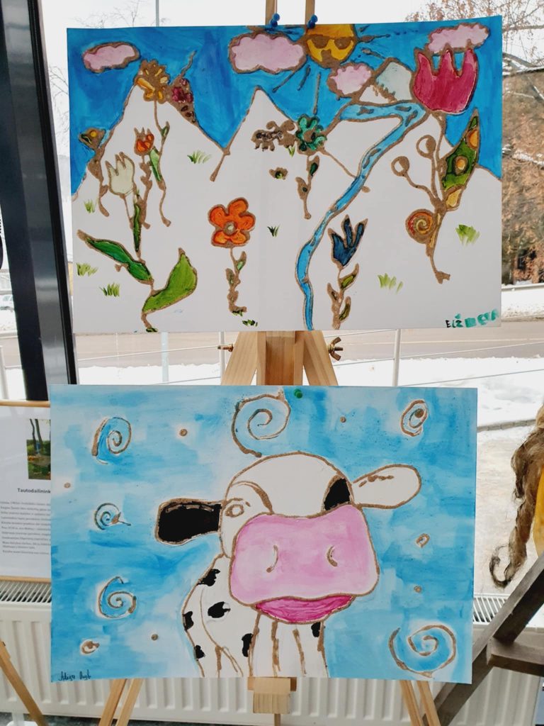 Two colorful artworks displayed on easels, featuring a landscape with mountains, flowers, and a river in the top painting, and a whimsical cow against a blue background in the bottom painting.