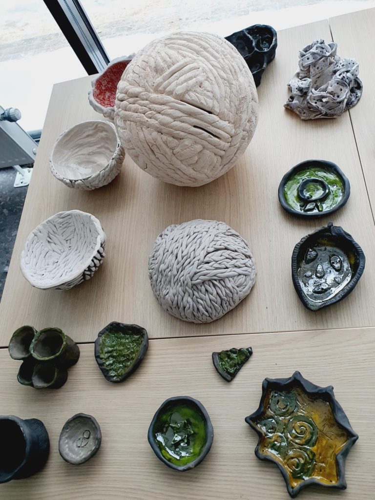 Handcrafted ceramic art pieces displayed on a wooden table, featuring a large textured spherical bowl, various small bowls in different shapes and colors, and intricate decorative plates with glossy finishes. The arrangement showcases a blend of natural tones and artistic designs, highlighting the craftsmanship of pottery.