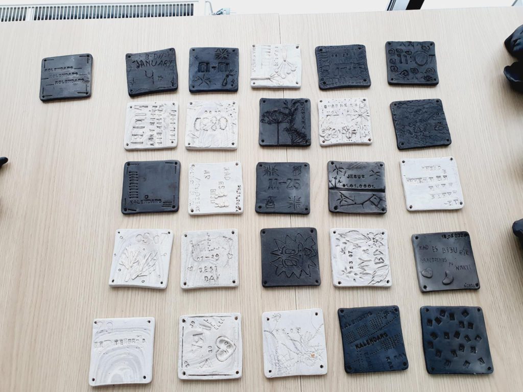 Collection of handmade clay tiles featuring various designs and inscriptions, displayed on a wooden table. The tiles include dates, names, and decorative patterns, showcasing artistic creativity and personalization.