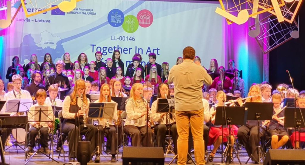 A vibrant performance scene featuring a diverse choir and musicians on stage, with a conductor leading the ensemble. The backdrop displays the event title "Together In Art" along with colorful lighting and hanging musical note decorations, highlighting a cultural collaboration between Latvia and Lithuania.