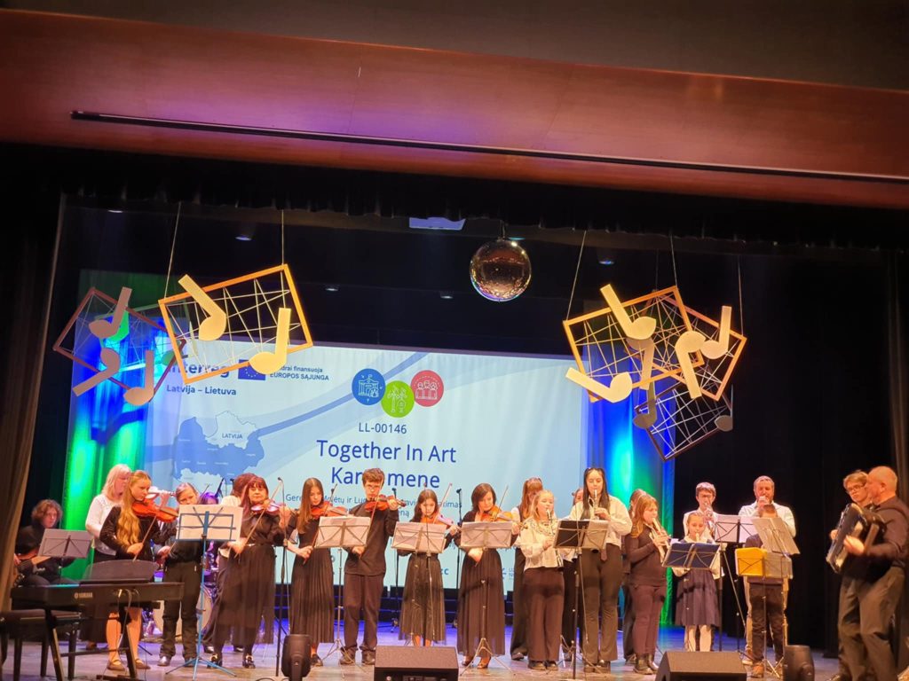 Students performing on stage during the "Together In Art" event, showcasing musical talents with violins and other instruments, surrounded by colorful music-themed decorations. A backdrop features the event title and logos representing Latvia and Lithuania.