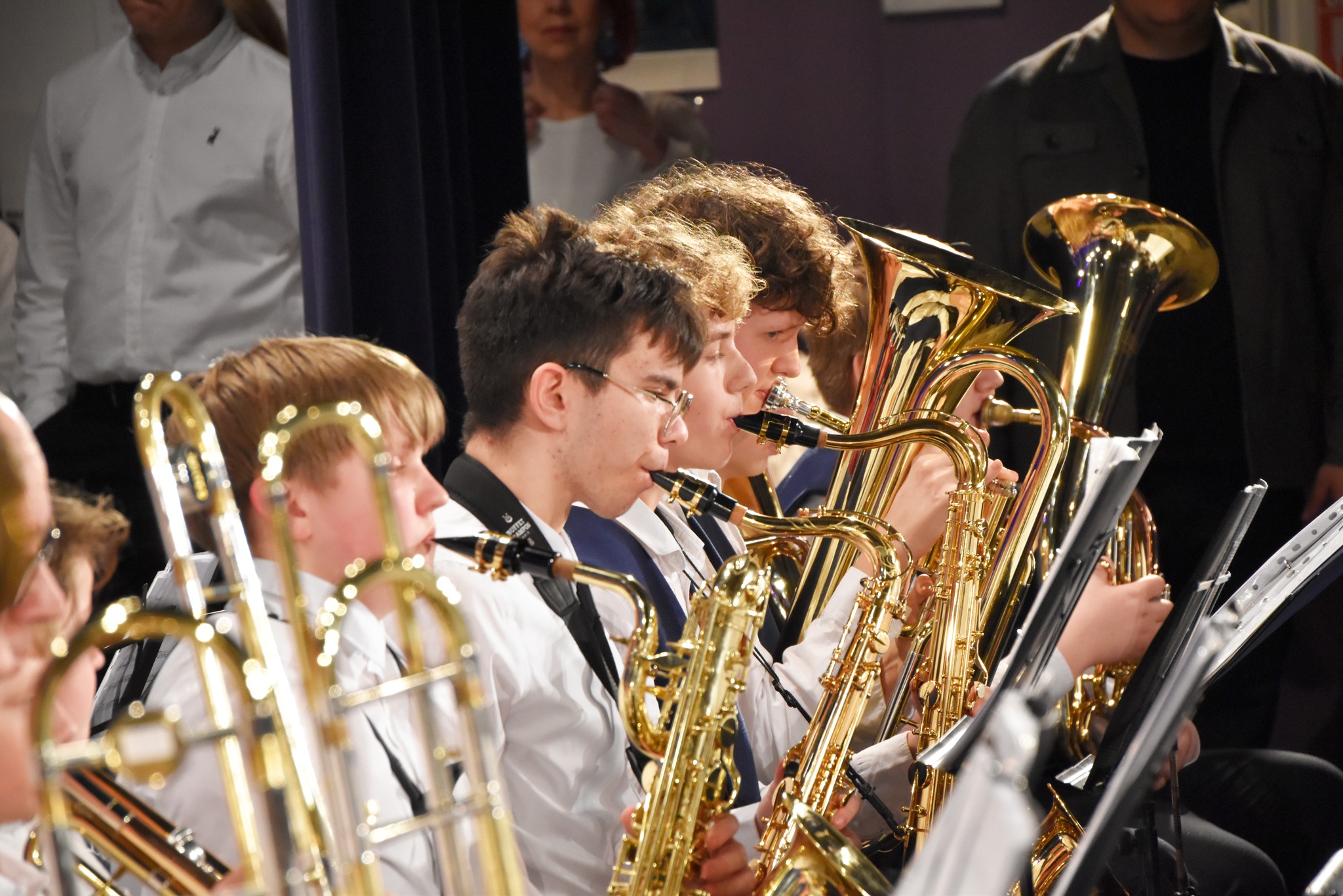 Young musicians performing in a concert, focusing on several players with brass instruments, including tubas and euphoniums, dressed in formal attire. The image captures the dedication and concentration of the band members during their performance.