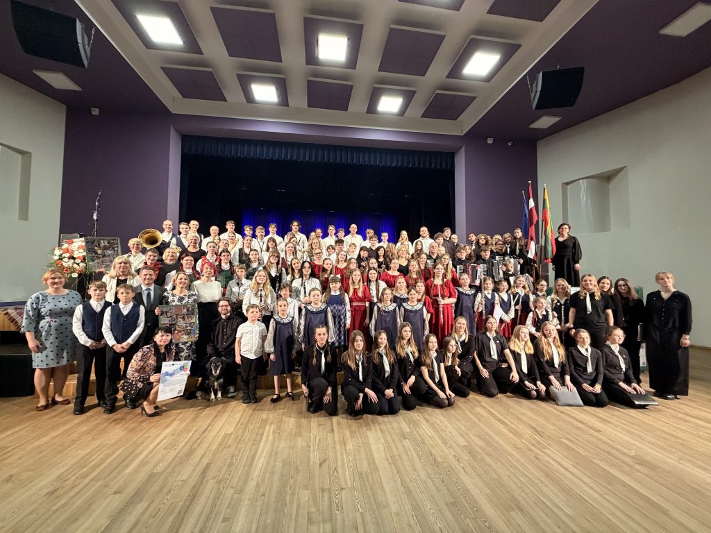 Group photo of a large choir and orchestra performing in a concert hall, featuring a diverse ensemble of children and adults dressed in formal attire and traditional costumes, with flags in the background and floral arrangements, showcasing community engagement in the arts.