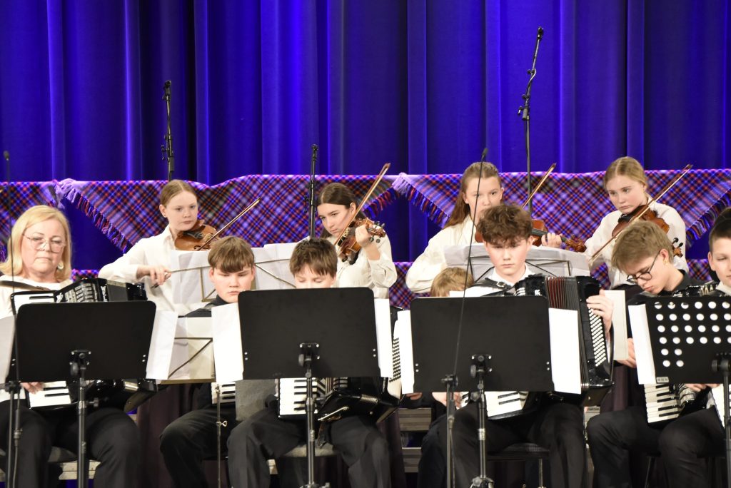 A group of young musicians performing on stage, featuring children playing violins and accordions, dressed in formal attire, with a colorful backdrop and stage lighting.