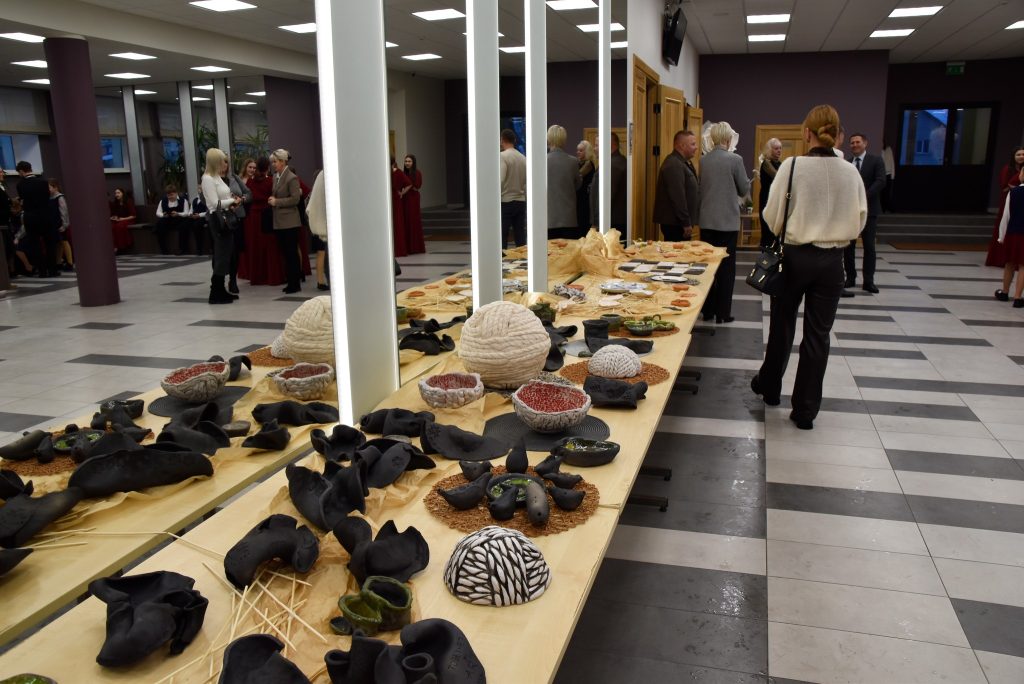 Art exhibition showcasing unique ceramic and textile art pieces on display in a spacious gallery. The scene features a long table adorned with various handcrafted items, including bowls and sculptures, while attendees engage in conversation in the background. The modern interior is well-lit, highlighting the intricate details of the artwork.