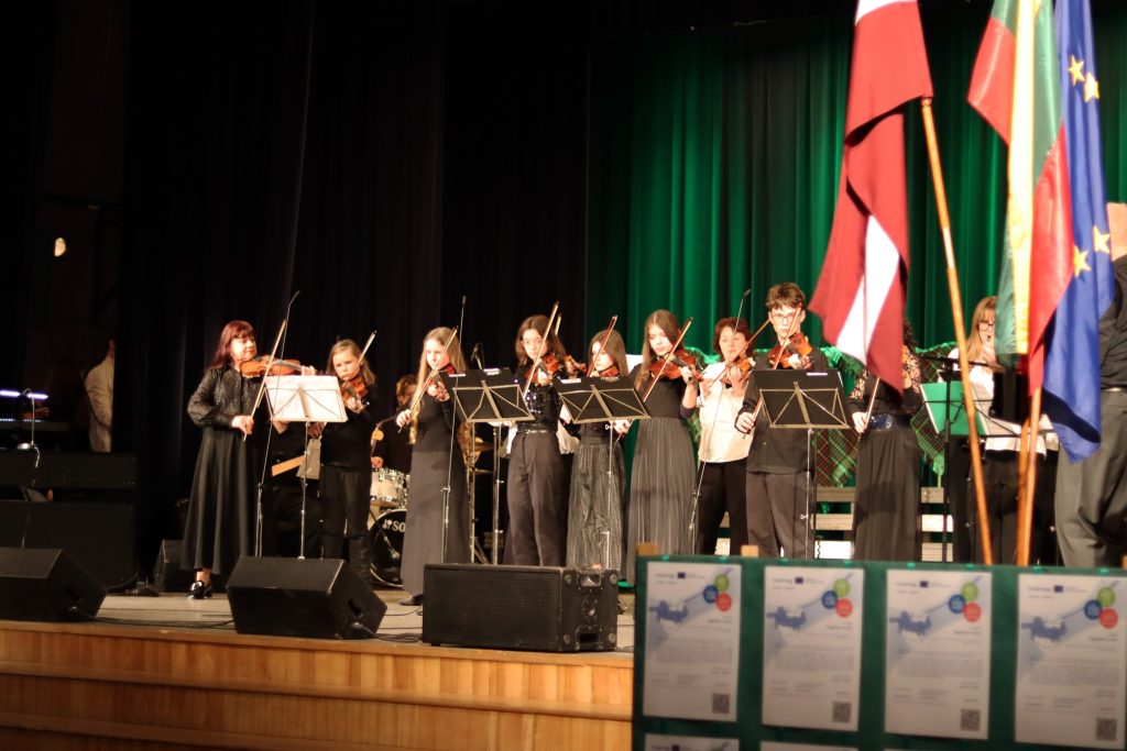 Young musicians performing a violin ensemble on stage, dressed in black attire, with flags in the background and musical instruments visible. The event showcases talent and cultural diversity, highlighting a live music performance in a festive setting.