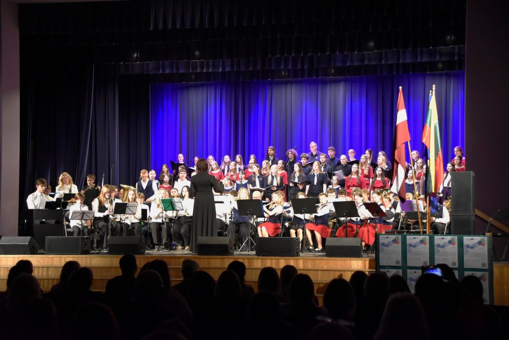A large choir and band perform on stage at a concert, featuring musicians in formal attire with a conductor leading from the front. The backdrop is illuminated with blue lighting, and flags from different countries are displayed. The audience is visible in the foreground, enjoying the performance.