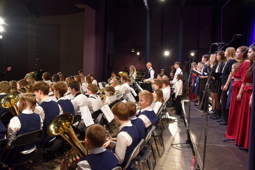 A school concert featuring a large ensemble of musicians in formal attire, including brass and woodwind instruments, performing on stage. A choir is positioned to the right, with a conductor leading the performance. Bright stage lights illuminate the scene, creating a vibrant atmosphere. Music sheets are visible on stands in front of the musicians.