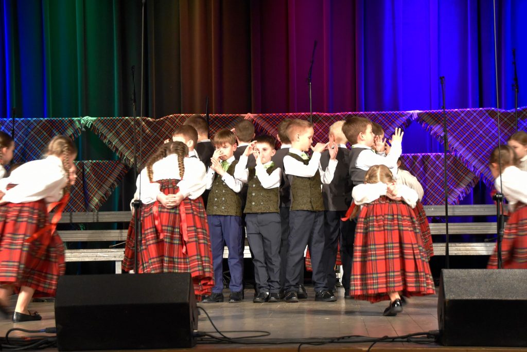 Children performing on stage in traditional attire, featuring plaid skirts and vests, during a school event. Colorful stage lighting enhances the festive atmosphere as they engage in a lively group performance.