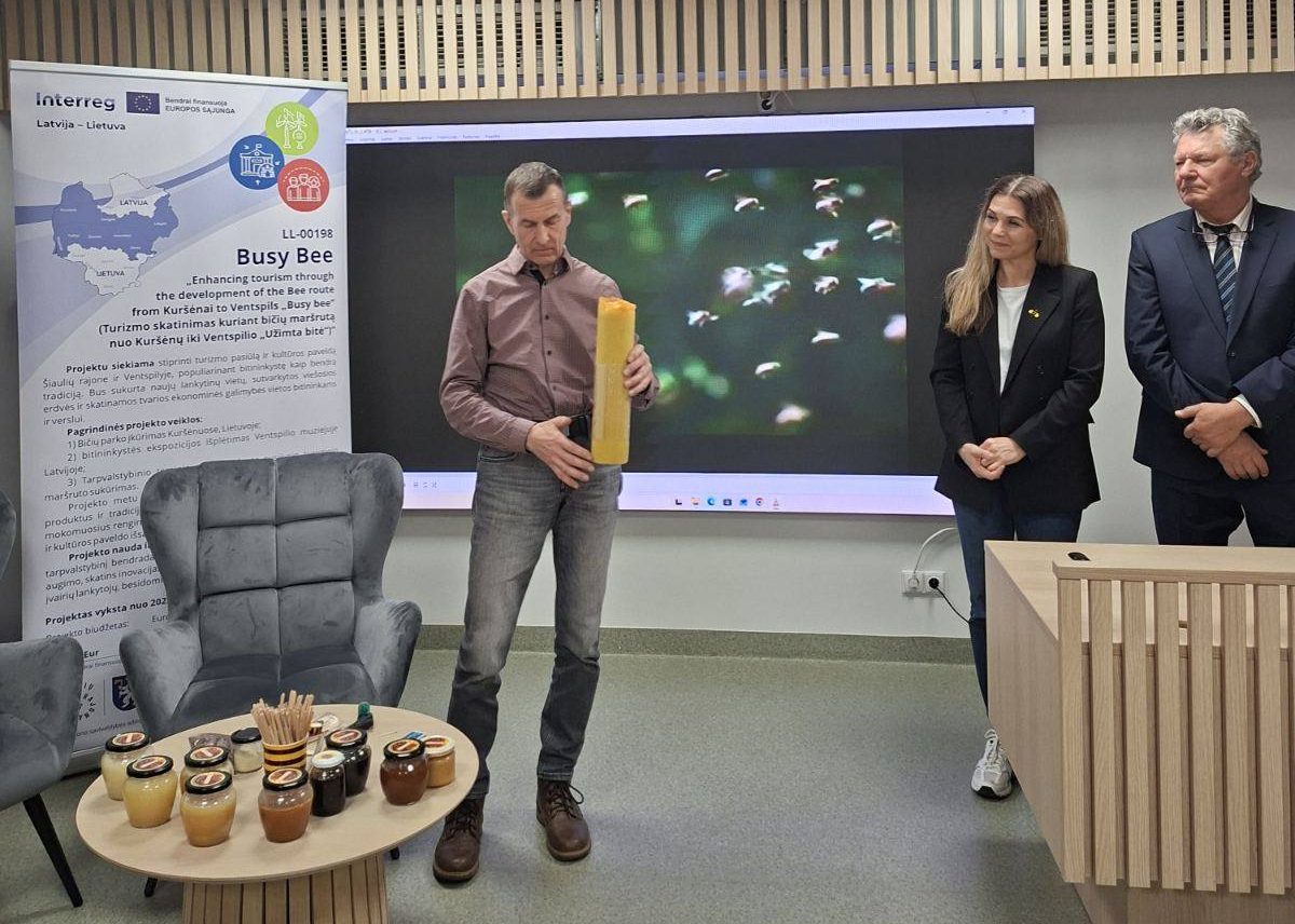 Presentation event showcasing honey products, featuring a speaker holding a beeswax candle in front of a screen displaying bees. Attendees include two individuals, one in a suit and another in casual attire, with a table displaying various jars of honey and wooden sticks for tasting. The backdrop includes an informational banner about the Busy Bee project, promoting local honey production and tourism in the Latvia-Lithuania region. Presentation event showcasing honey products, featuring a speaker holding a beeswax candle in front of a screen displaying bees. Attendees include two individuals, one in a suit and another in casual attire, with a table displaying various jars of honey and wooden sticks for tasting. The backdrop includes an informational banner about the Busy Bee project, promoting local honey production and tourism in the Latvia-Lithuania region.