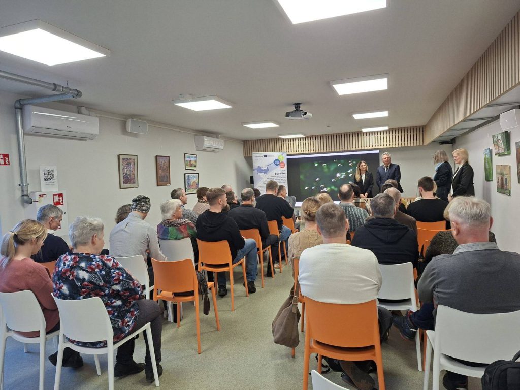Audience attending a presentation in a well-lit conference room, featuring a panel of speakers at the front and various artworks displayed on the walls. The attendees are seated in orange and white chairs, engaged in the discussion. Audience attending a presentation in a well-lit conference room, featuring a panel of speakers at the front and various artworks displayed on the walls. The attendees are seated in orange and white chairs, engaged in the discussion.