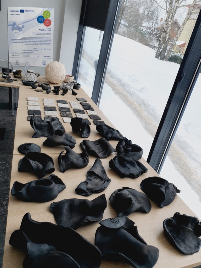Exhibition display featuring various black ceramic artworks on a wooden table, with a backdrop of a snowy landscape and a promotional poster for the "Together in Art" project. The pieces exhibit intricate designs and textures, showcasing contemporary ceramic artistry.