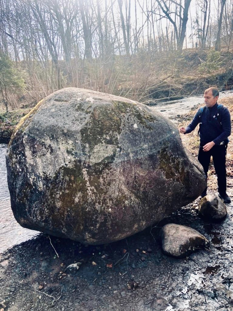 A person examining a large moss-covered boulder near a creek, surrounded by trees in a natural setting. The sunlight filters through the branches, highlighting the rocky terrain and the individual’s exploration of the geological feature.