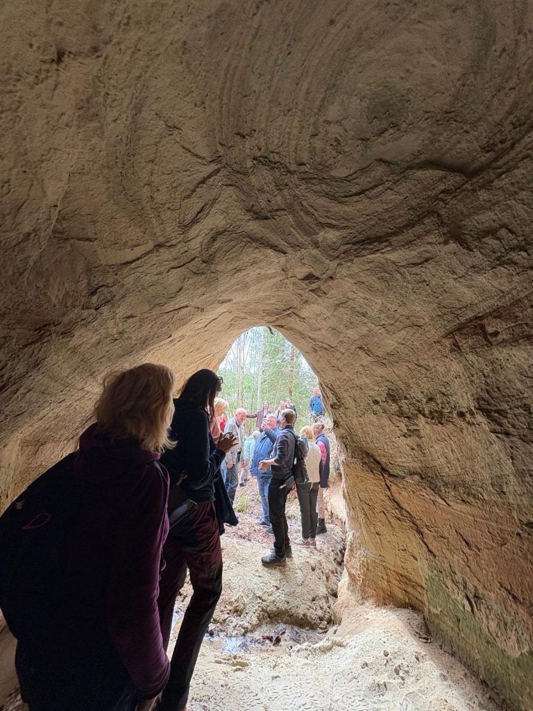 Group of people gathered in a cave-like structure surrounded by natural rock formations, with a view of trees in the background. The image captures the exploration and adventure atmosphere of a hiking or nature excursion.