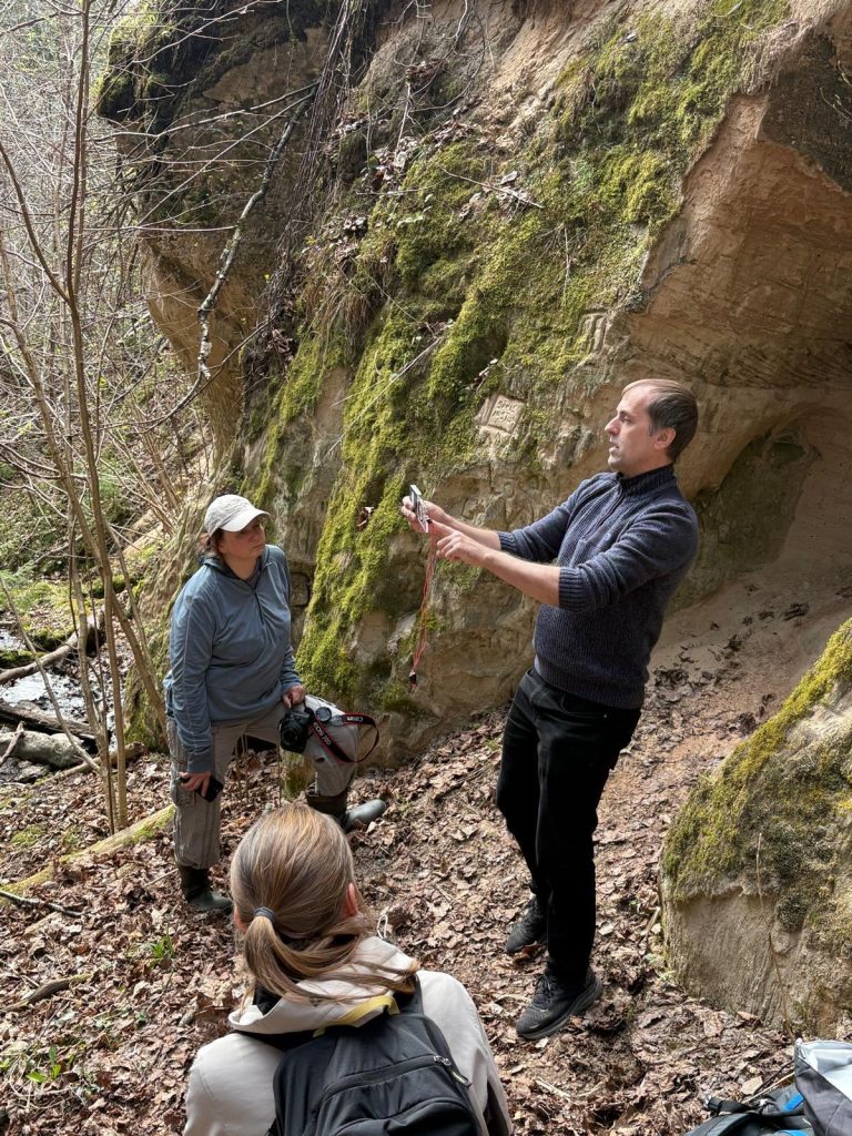Group of outdoor enthusiasts engaged in a nature education session in a forested area, with one person demonstrating a technique while others listen attentively, surrounded by moss-covered rocks and fallen leaves.