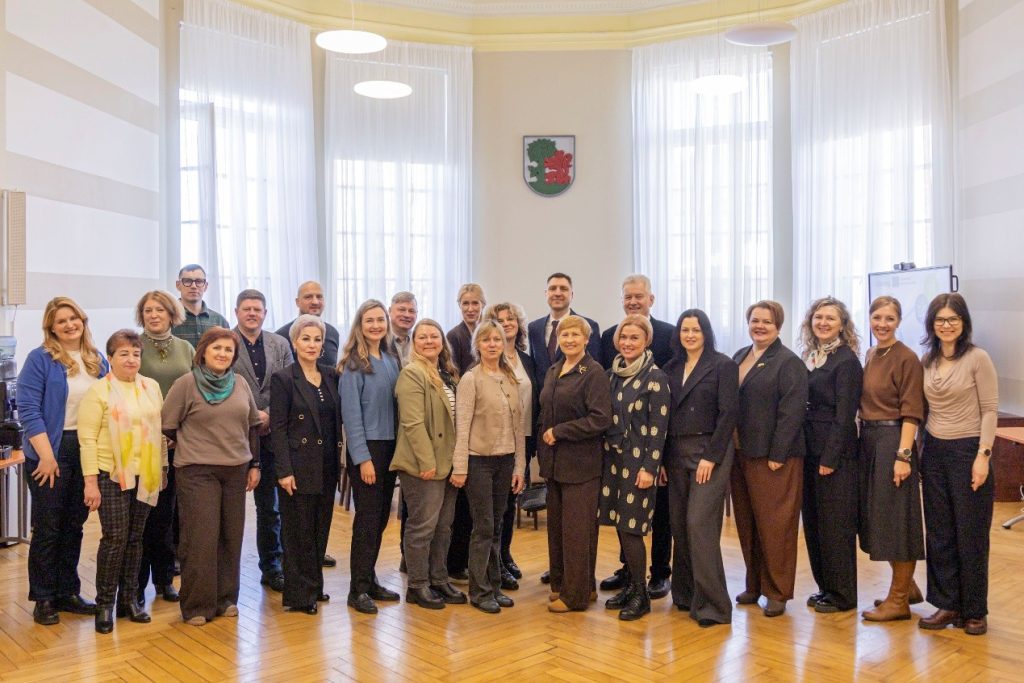 Group photo of 24 professionals posing together in a well-lit conference room with large windows and a coat of arms on the wall. The individuals are dressed in business attire, showcasing a diverse mix of styles and colors, representing a collaborative meeting or event.