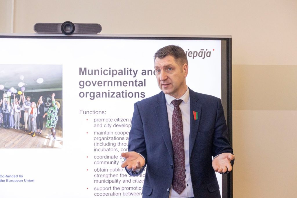 A speaker presenting on the roles of municipalities and governmental organizations, highlighting functions such as promoting citizen engagement, coordinating community initiatives, and fostering cooperation between municipalities and citizens. The presentation includes a visual of a group celebrating with balloons in the background.